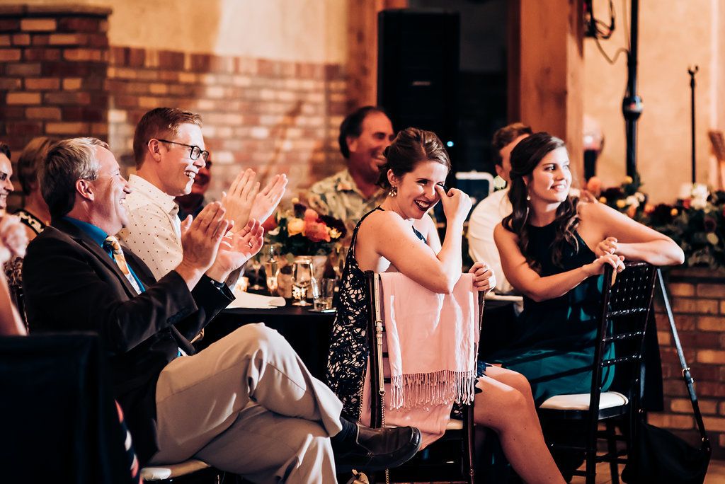 Wedding guests applaud and celebrate during an evening reception at a rustic brick venue with string lights.