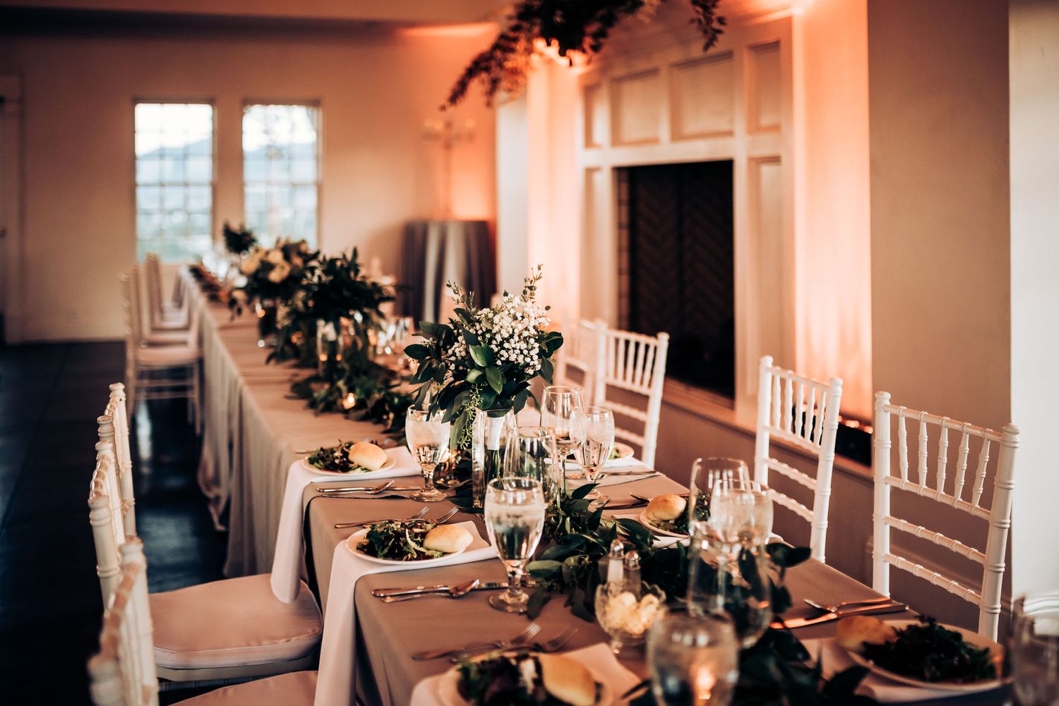 Long banquet table set for wedding reception with white chairs and floral centerpieces.