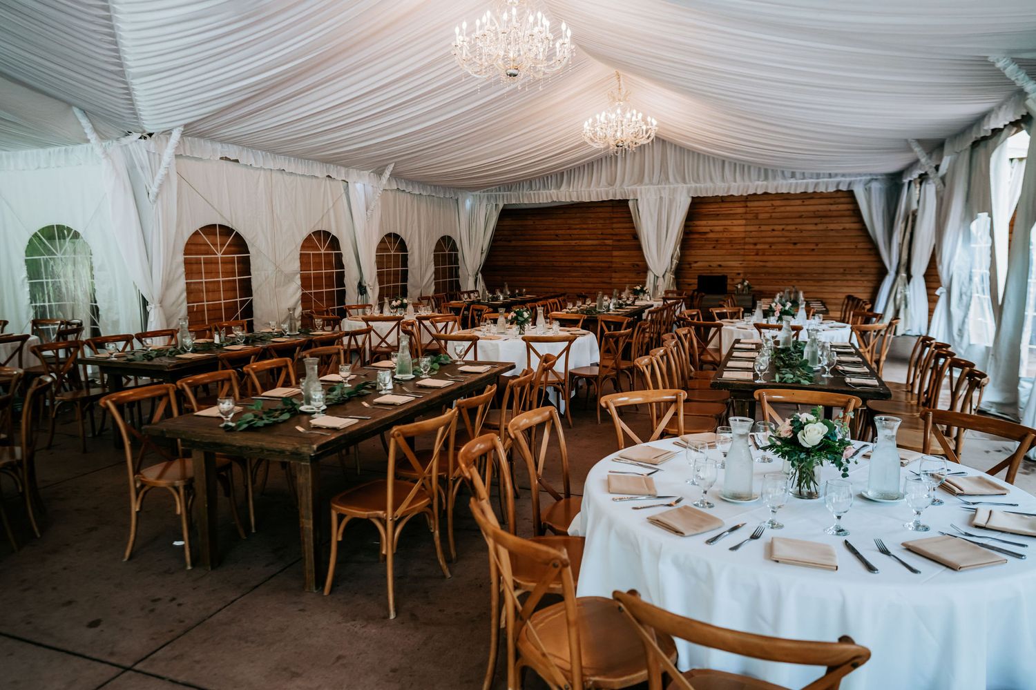 Elegant wedding reception setup with round tables and wooden chairs inside draped white tent.