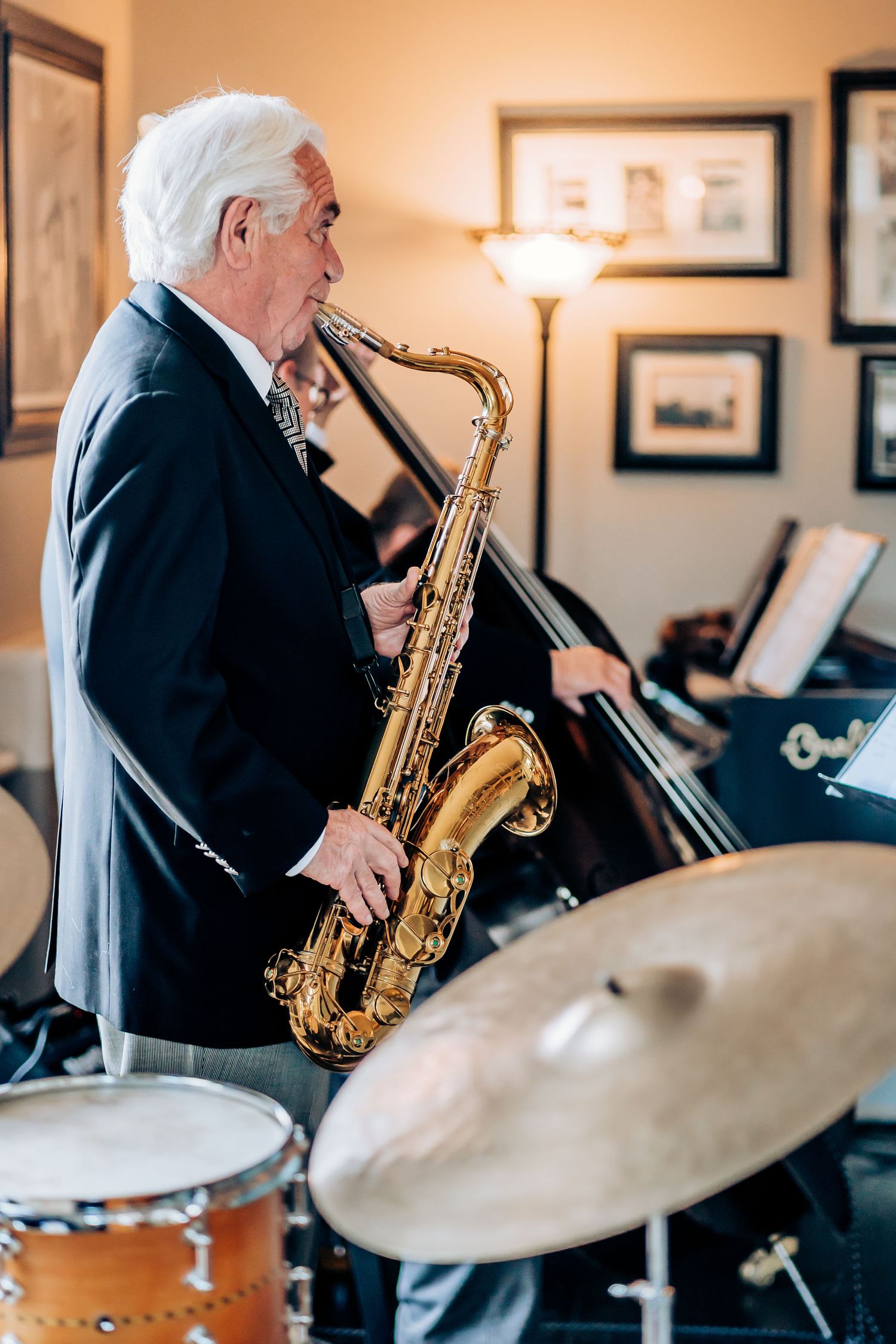 Senior musician in suit plays saxophone next to drum set in elegant room with framed artwork.