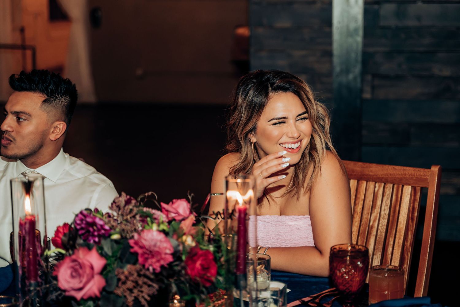 A guest at a wedding reception laughs at a table decorated with pink and purple flowers.