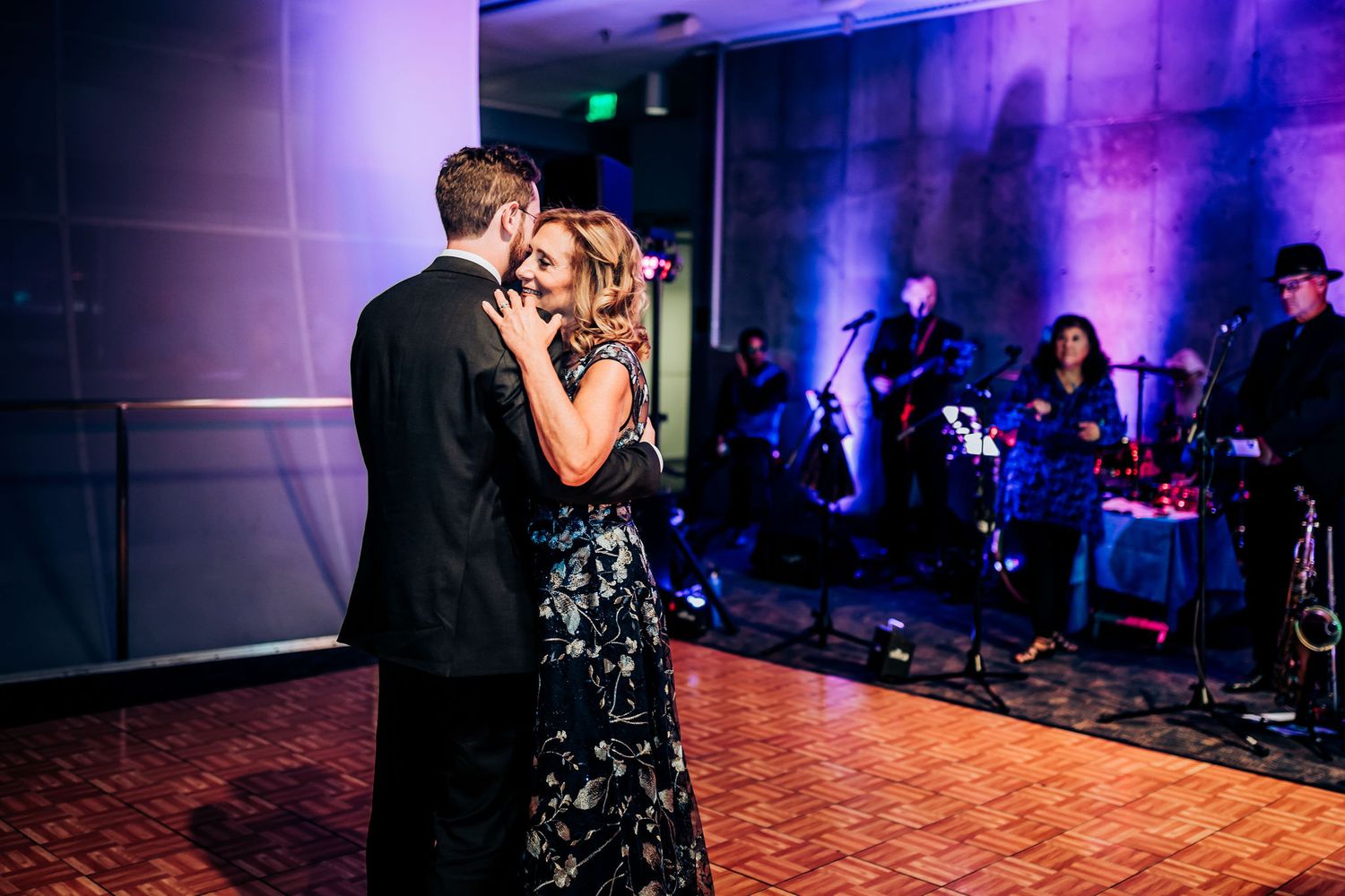 A couple dances intimately on the dance floor with purple mood lighting and guests in background.