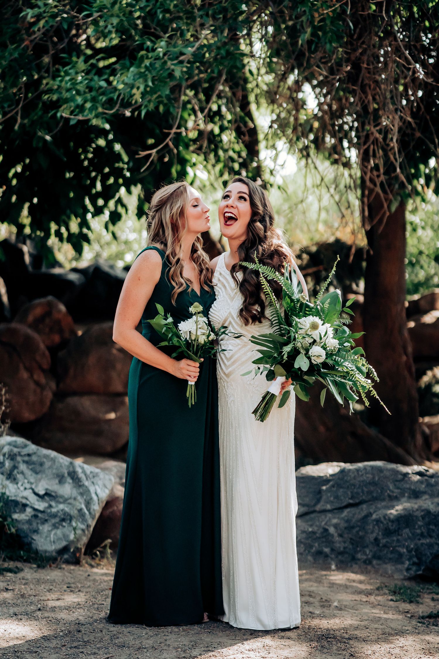 A joyful moment shared between wedding party members holding green and white bouquets outdoors.