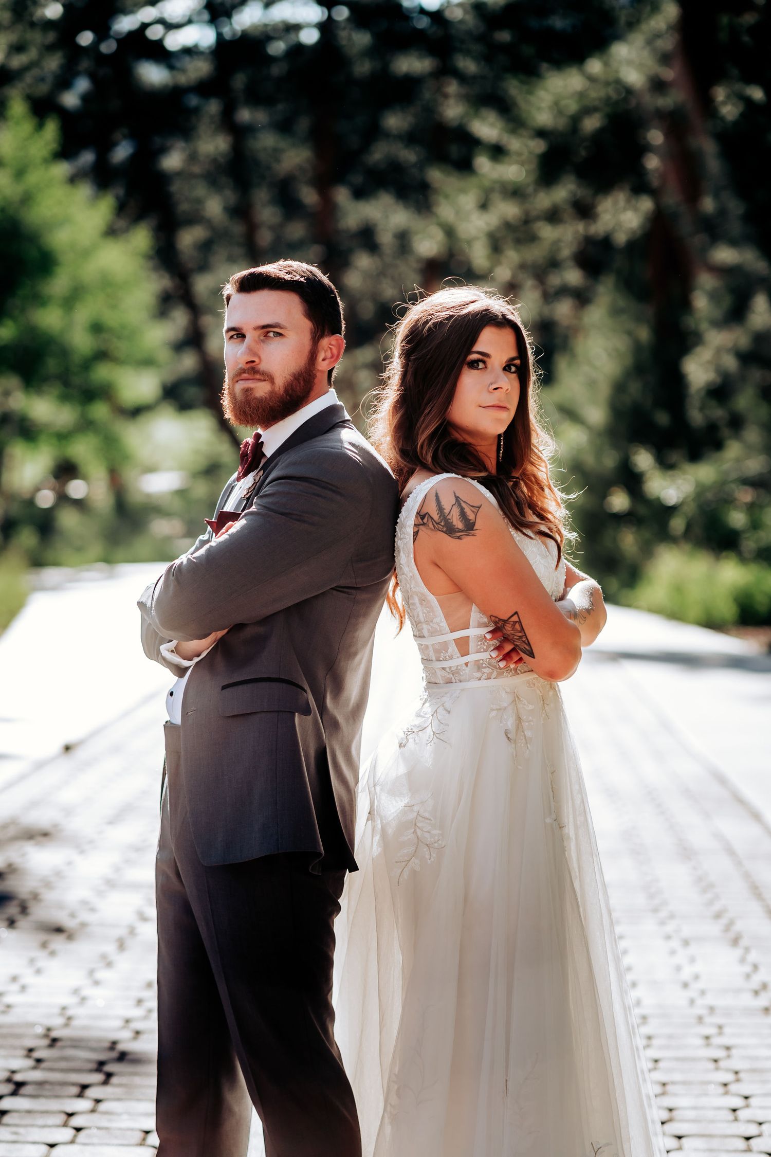 A bride in white and groom in dark suit pose back-to-back on a paved outdoor pathway.