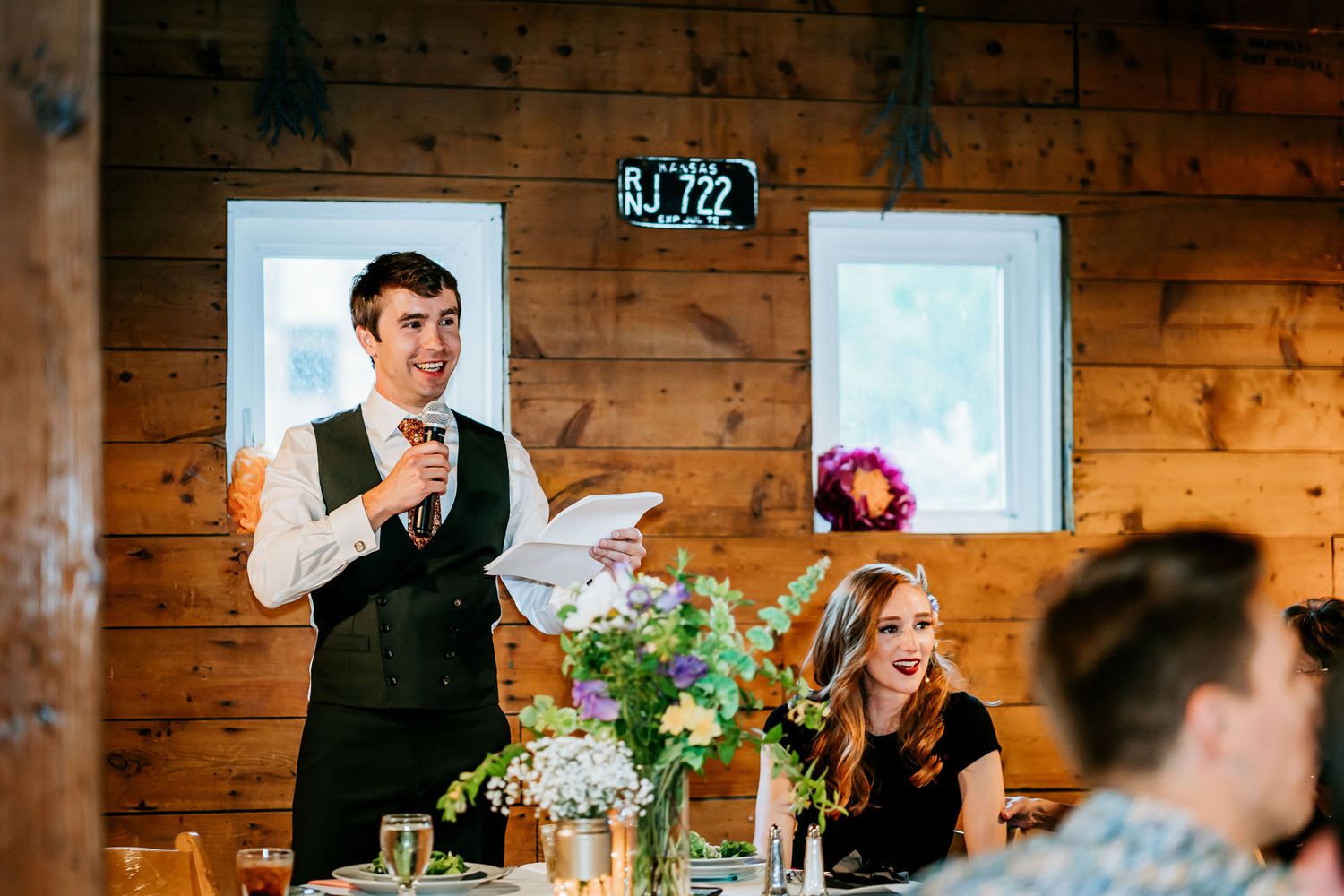 Wedding reception toast inside rustic wooden venue with string lights and floral centerpieces.