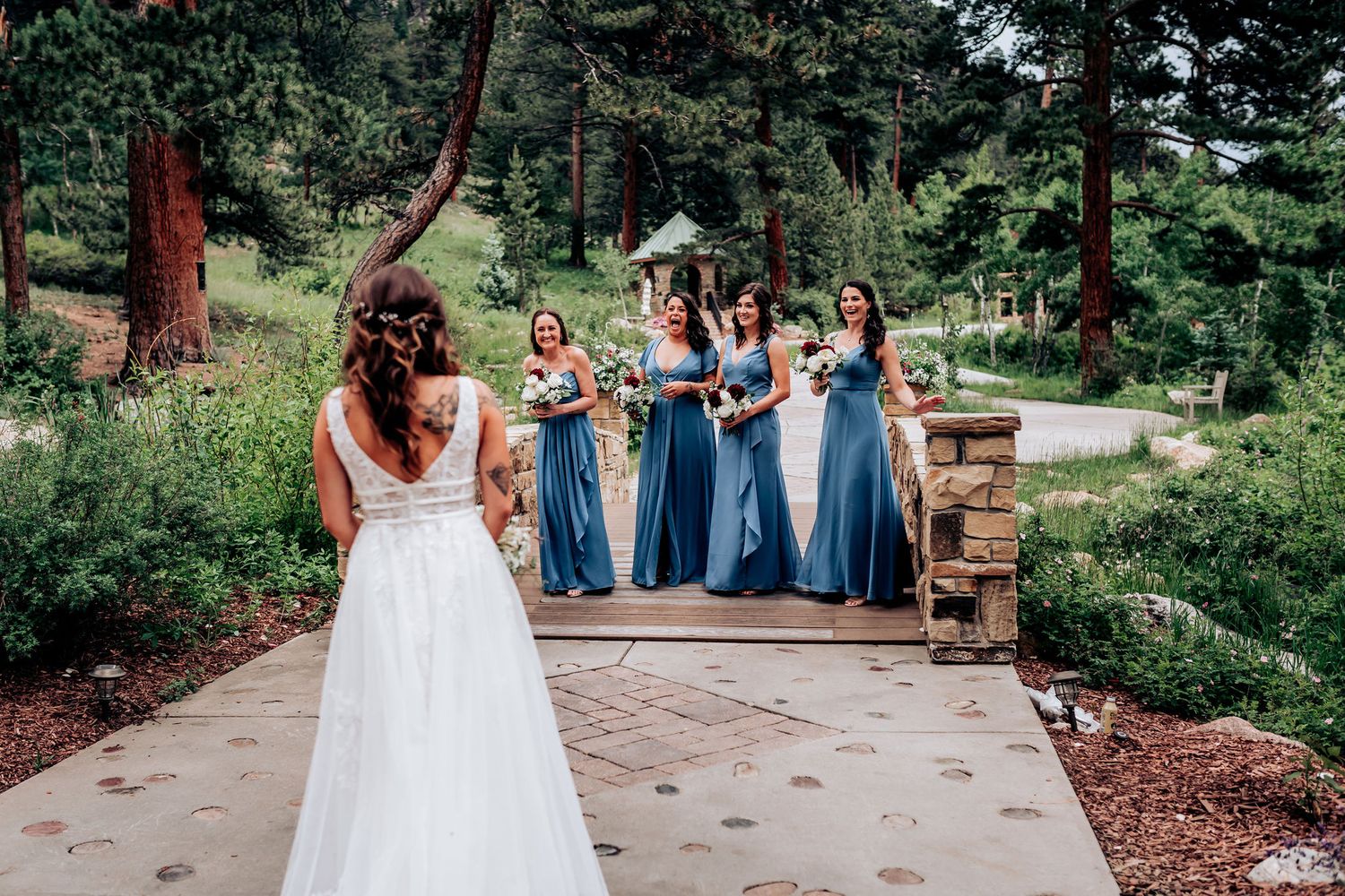 A wedding party in blue dresses gathers on a stone pathway surrounded by pine trees.