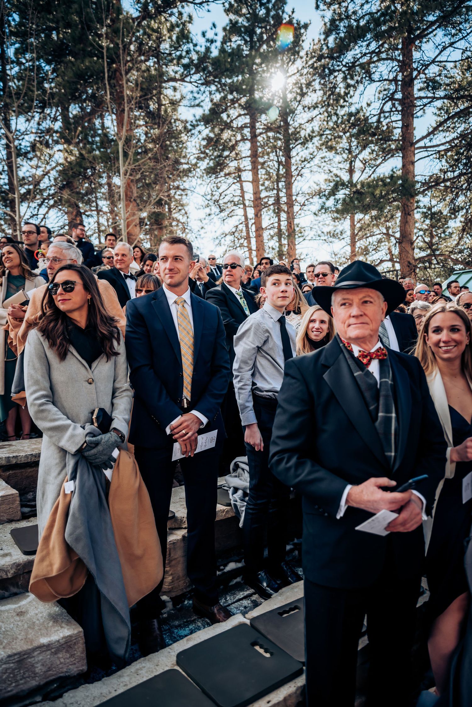 Formally dressed wedding guests standing among pine trees at mountain ceremony venue.