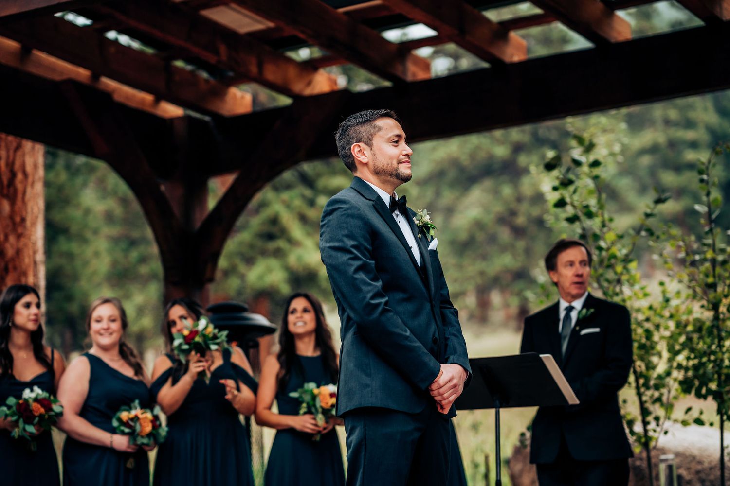 A wedding ceremony takes place under a wooden pergola with attendants in navy blue dresses.