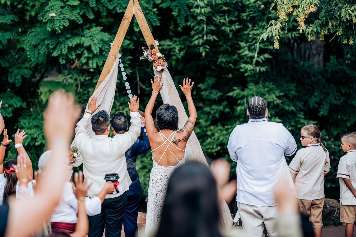 Wedding guests and attendees raise their arms in celebration under a wooden ceremony arch outdoors.