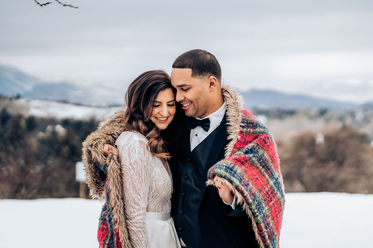Couple embraces under cozy blanket during winter mountain wedding photo session.