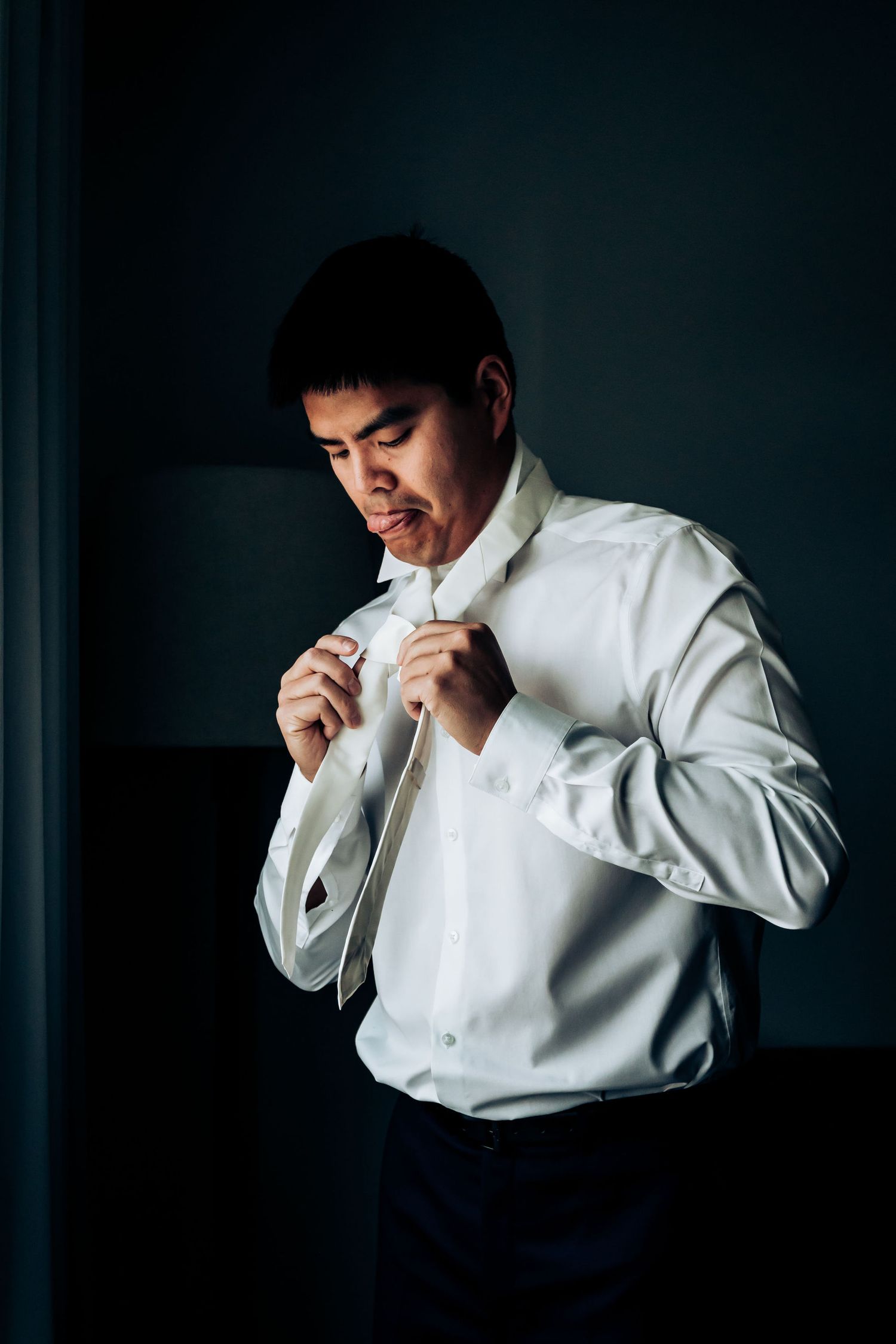 Groom adjusting white dress shirt collar in moody dramatic lighting.