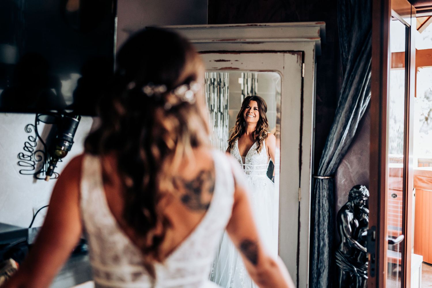 A bride in a white wedding dress looks at her reflection in an ornate mirror.