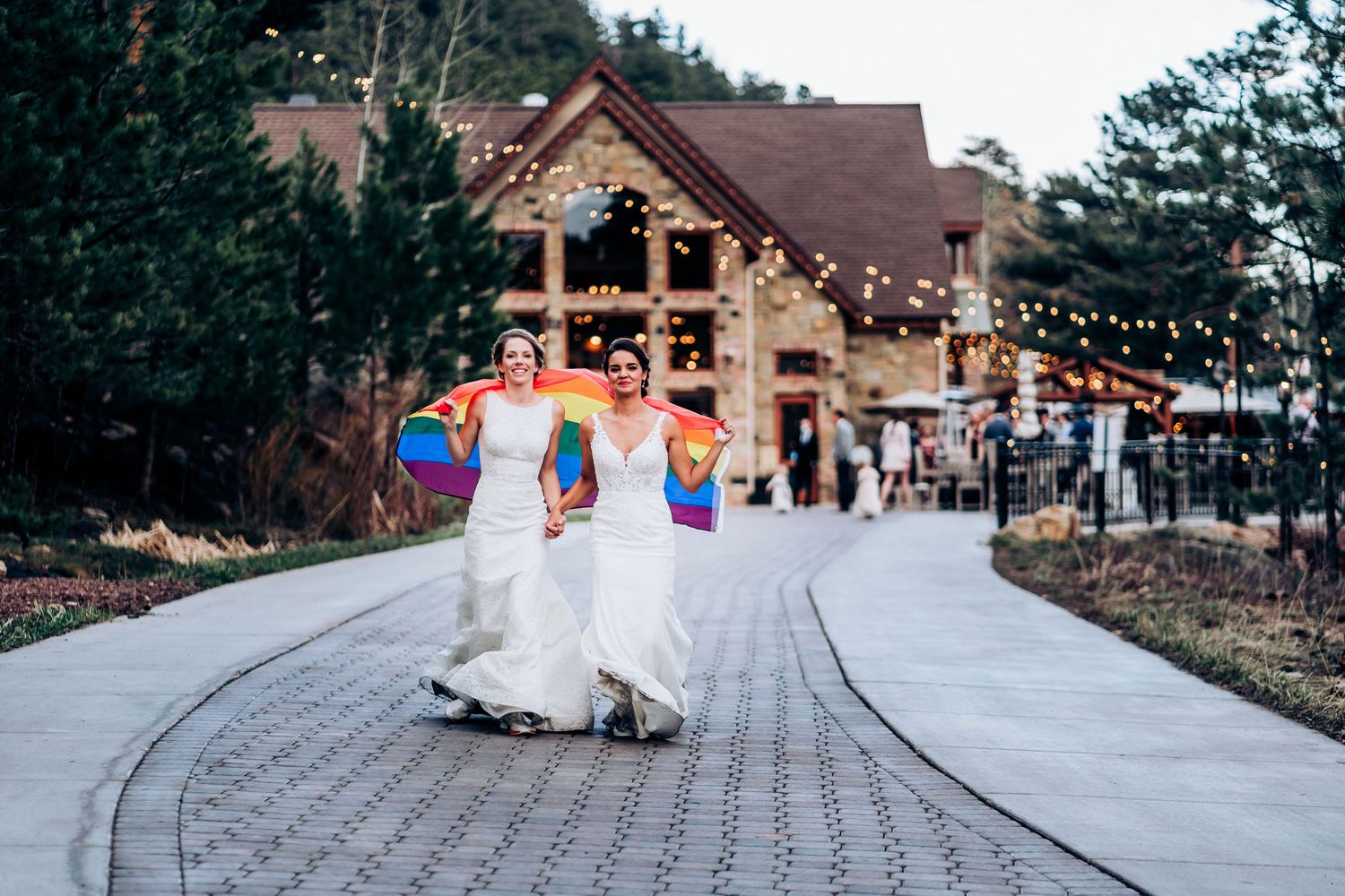 Two people in elegant white dresses dance together on a paved path leading to a rustic lodge decorated with string lights.