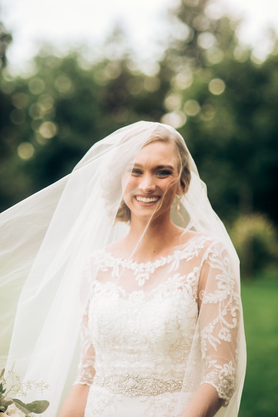 Joyful bride wearing lace gown and flowing veil smiles radiantly in outdoor garden setting.