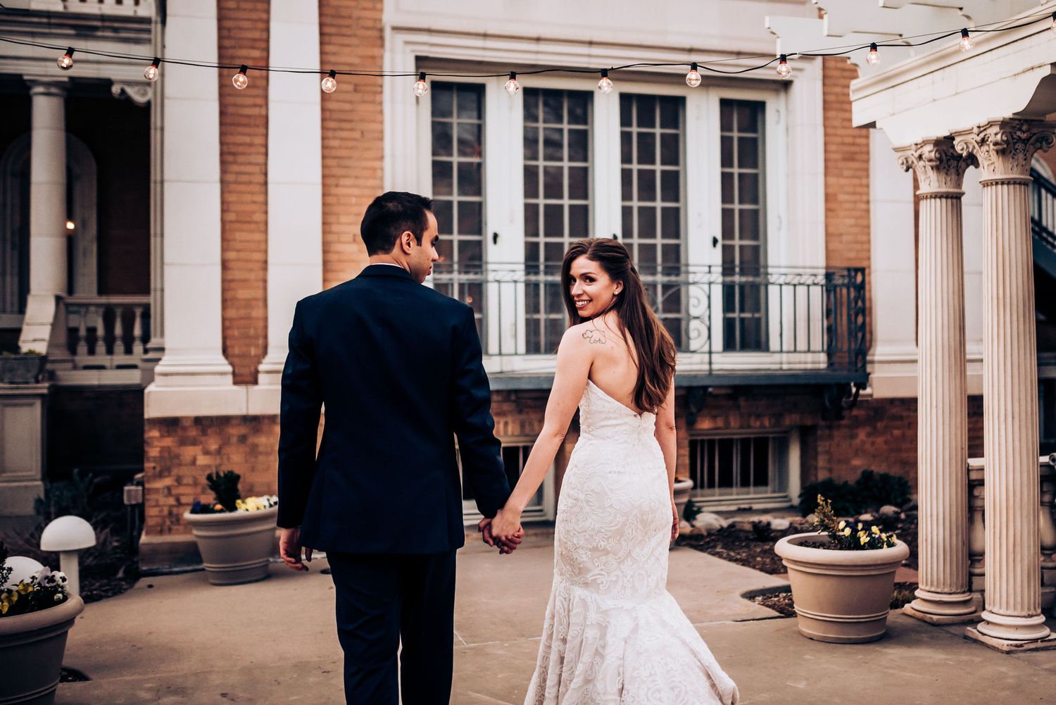 A couple walks hand in hand through a grand columned walkway with string lights overhead.