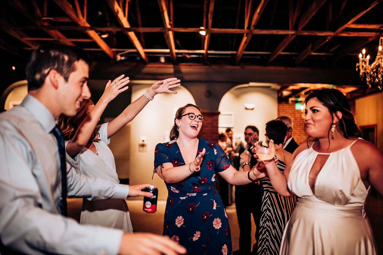 Wedding guests dance and celebrate at a reception in a rustic venue with exposed wooden beams and brick walls.
