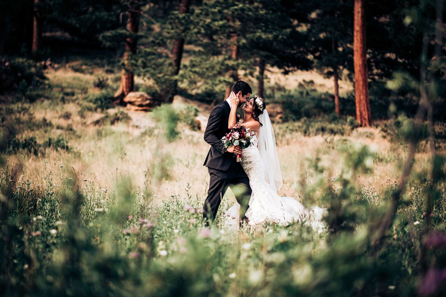 Wedding couple embraces in mountain meadow surrounded by wildflowers and pine trees.