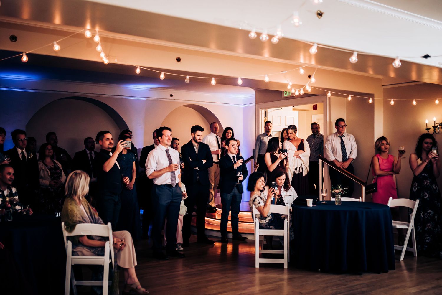 Wedding reception guests gather in a beautifully lit ballroom with string lights and archways.