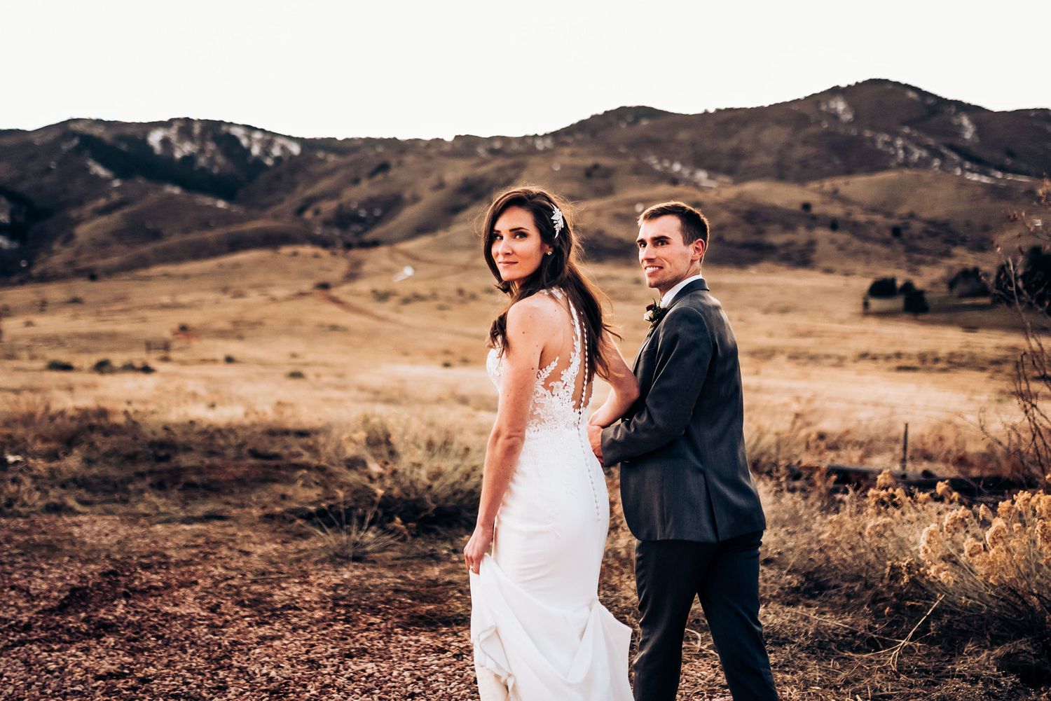 Elegant couple in wedding attire poses together on scenic mountain trail.