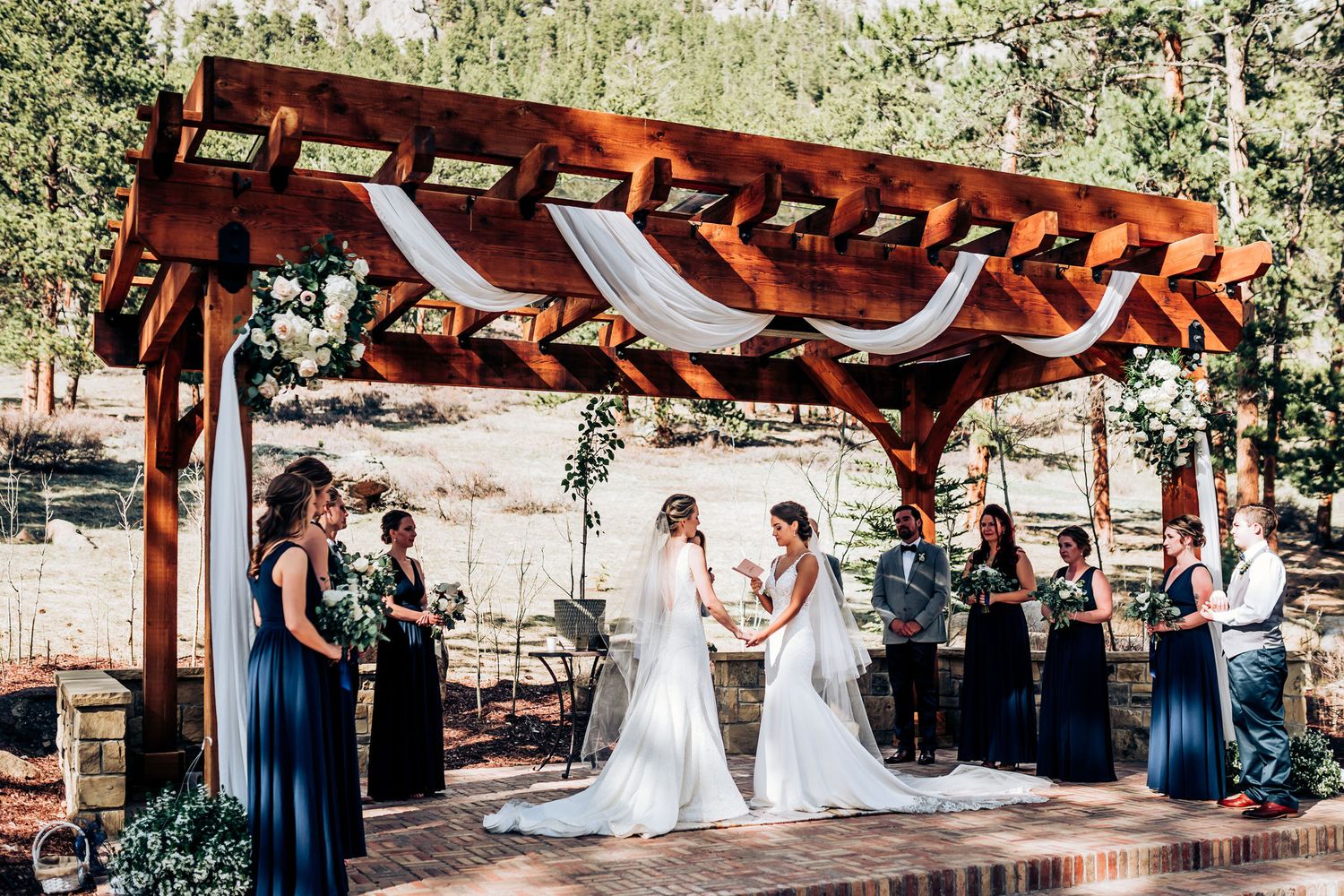 Wedding ceremony under rustic pergola with navy-clad attendants and mountain scenery.