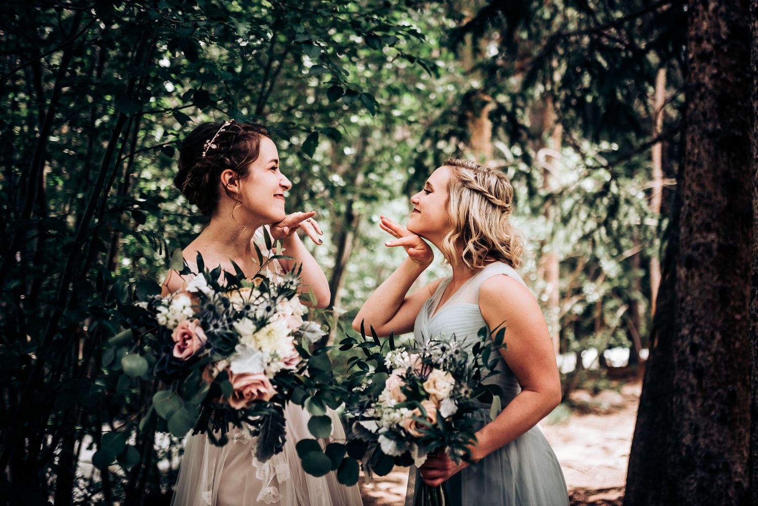 Two bridesmaids share joyful moment while holding floral bouquets in forest setting.