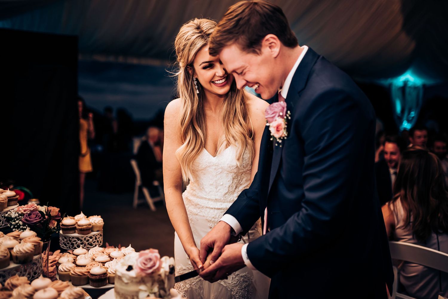 Newlyweds share a sweet moment while cutting their wedding cake together in a dimly lit reception tent.