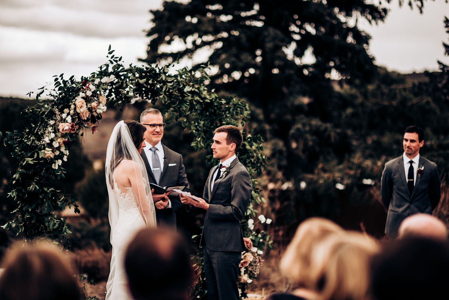 Wedding ceremony takes place under a natural floral arch with gathered guests in an outdoor setting.