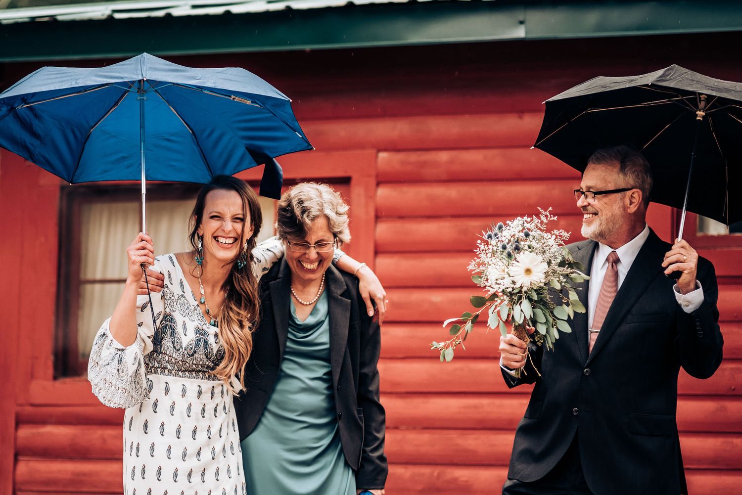 Wedding attendees with blue umbrellas pose in front of red cabin.