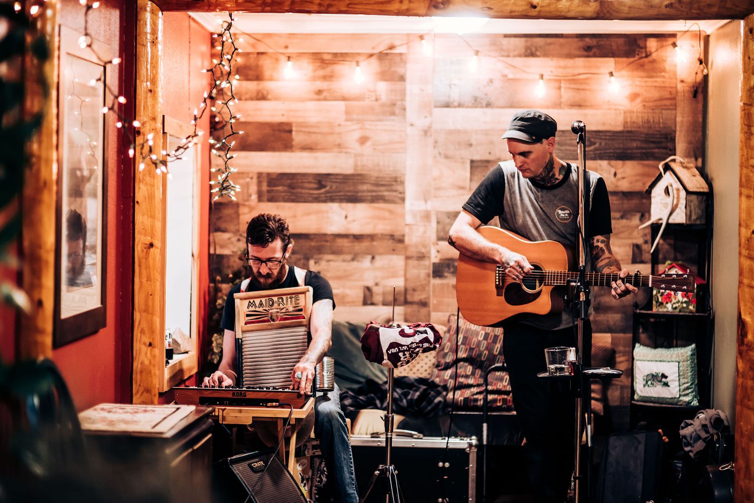 Musicians perform with acoustic guitar and keyboard in wood-paneled room with string lights.