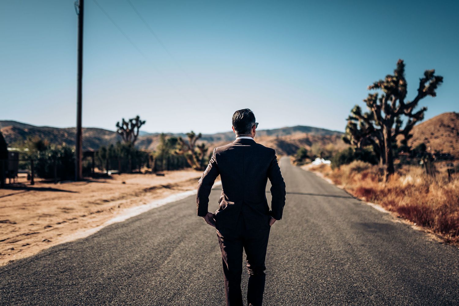 Person in formal suit walking alone on desert highway with Joshua trees.