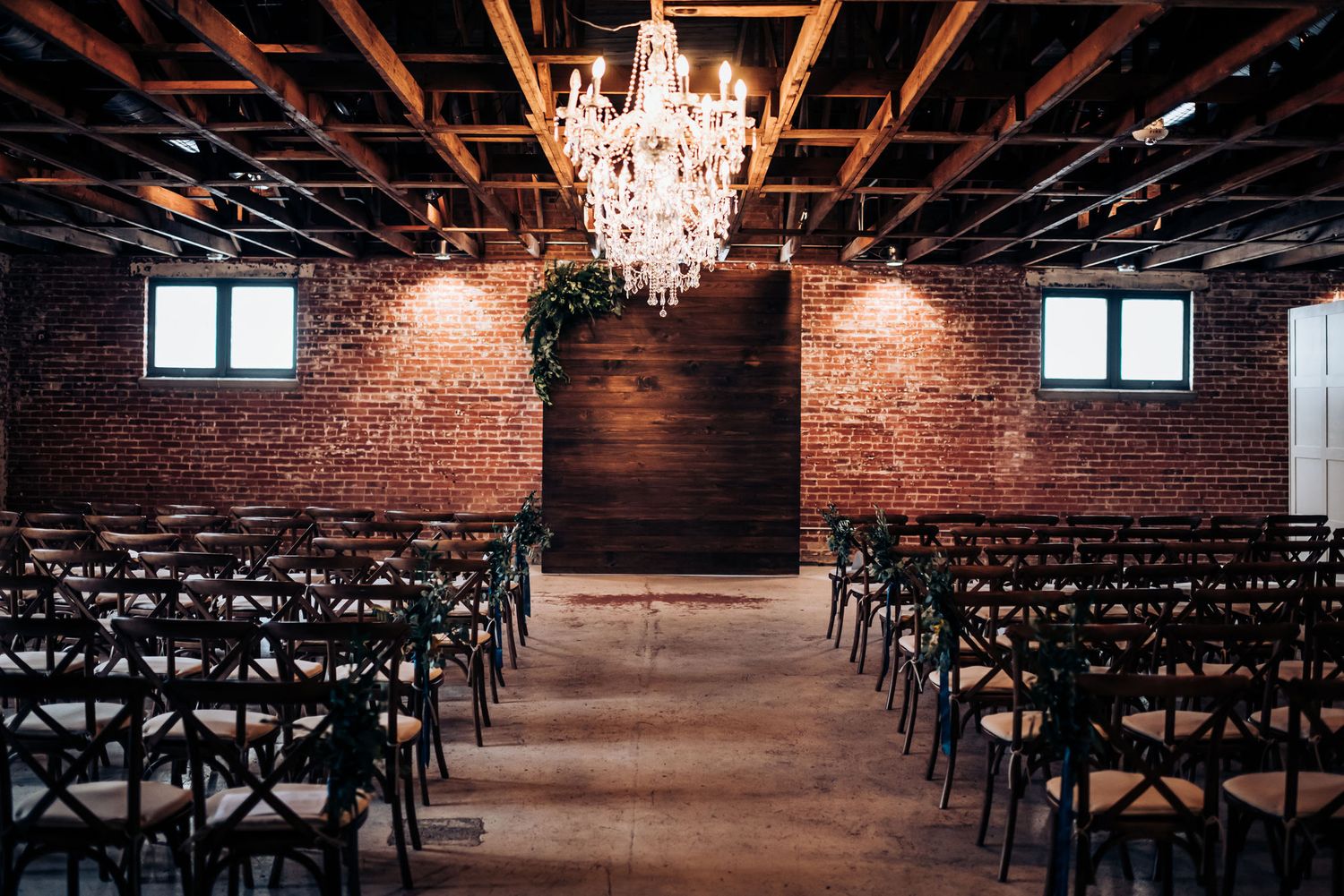 An elegant wedding ceremony space with exposed brick walls, crystal chandelier and dark wooden chairs.
