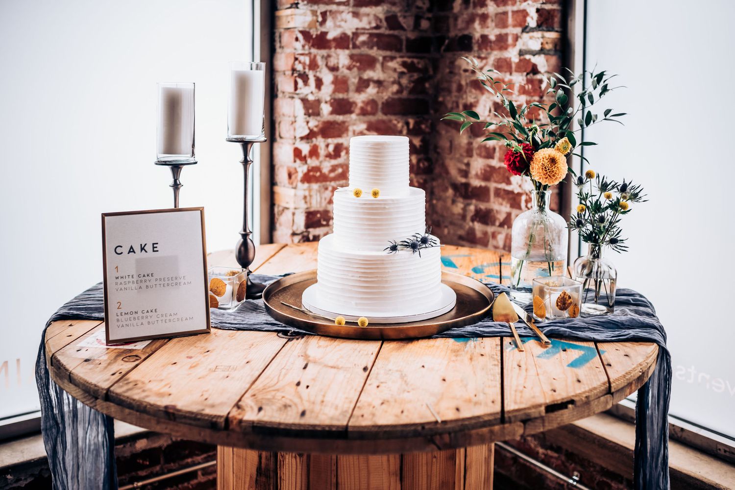 White tiered wedding cake displayed on wooden table with floral decorations against brick wall.