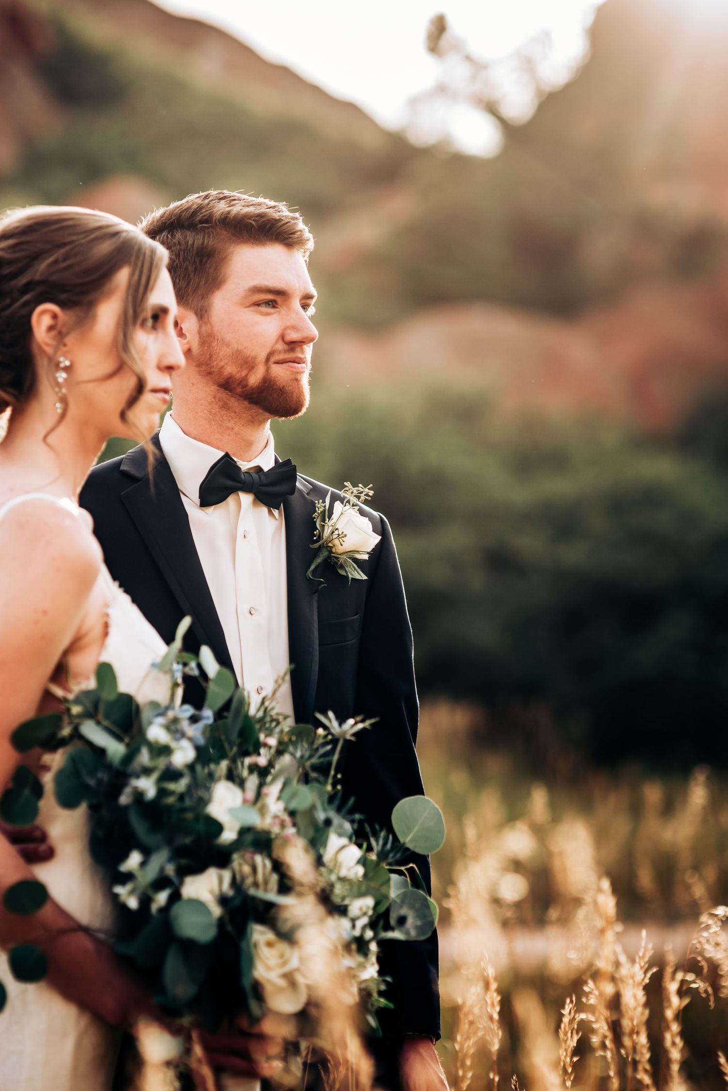Sunset wedding portrait with couple standing among dried grass and mountain backdrop.