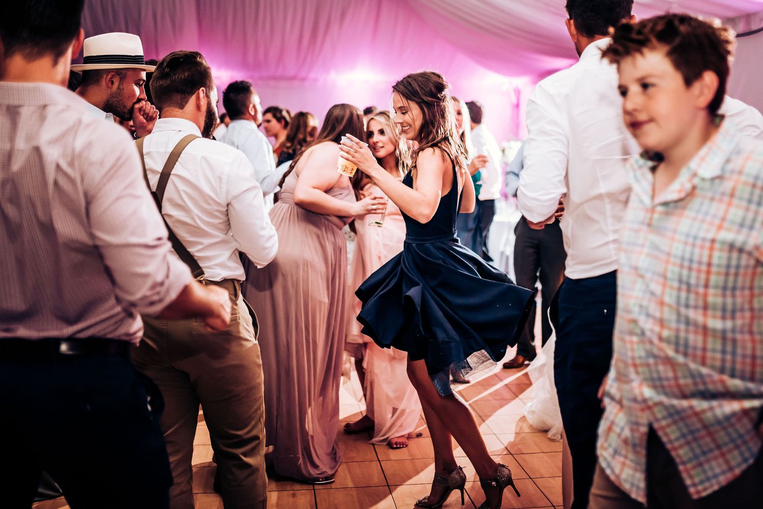 Party guests dancing energetically at a wedding reception in a pink-lit tent venue.