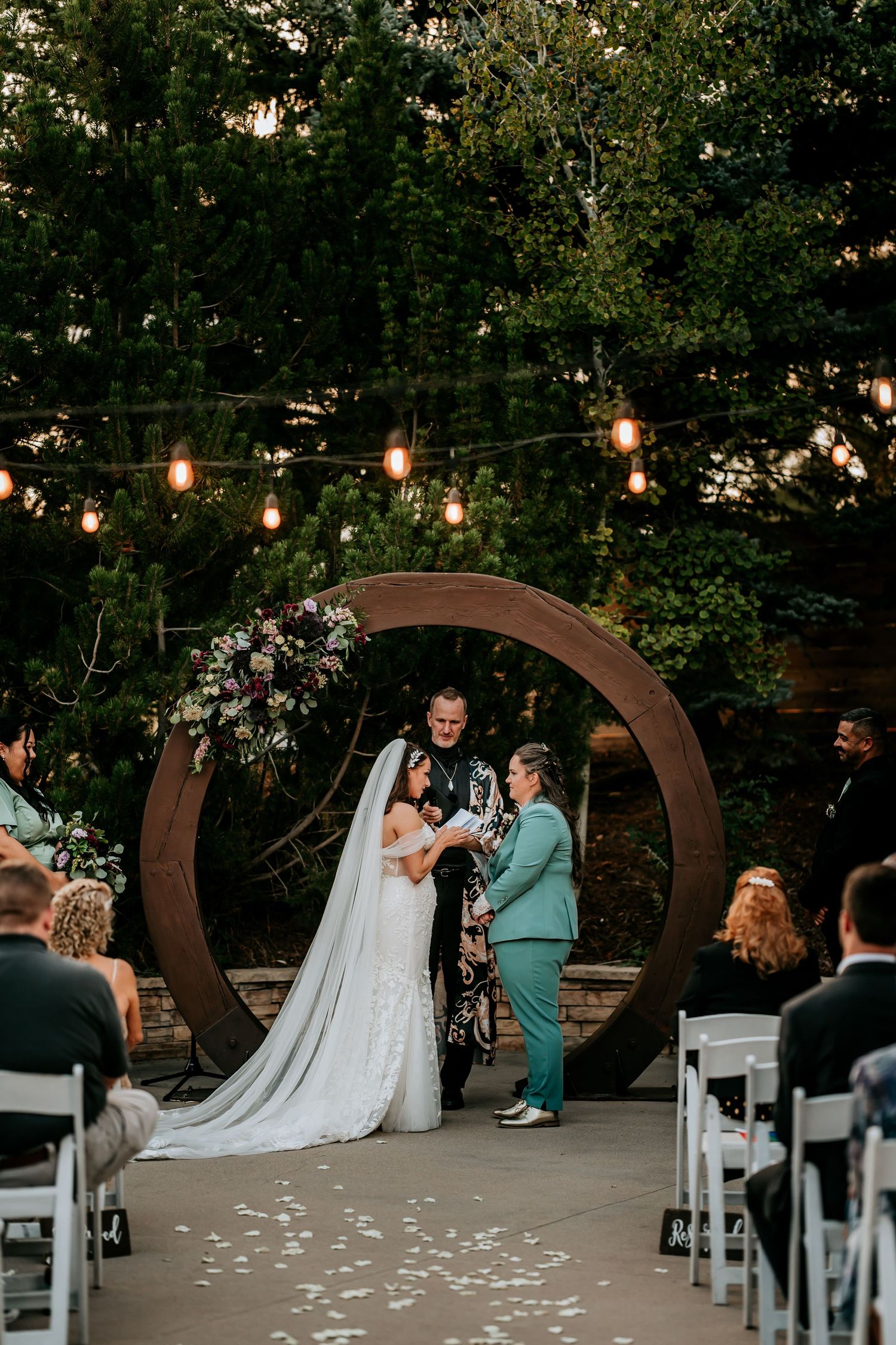 Outdoor wedding ceremony with couple at circular wooden arch adorned with flowers, string lights, and seated guests.