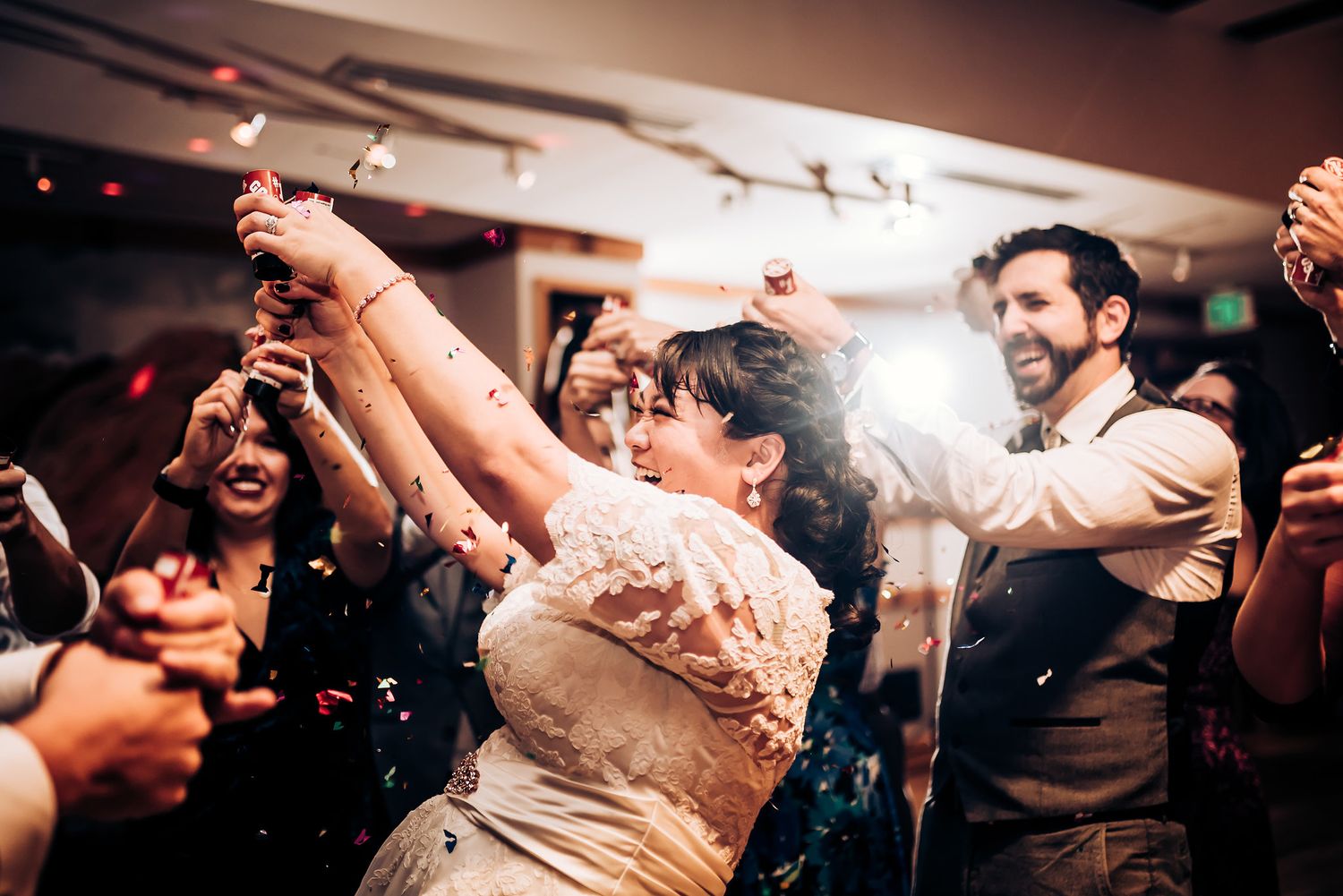 Wedding guests celebrate joyfully on the dance floor with raised arms during an indoor reception celebration.