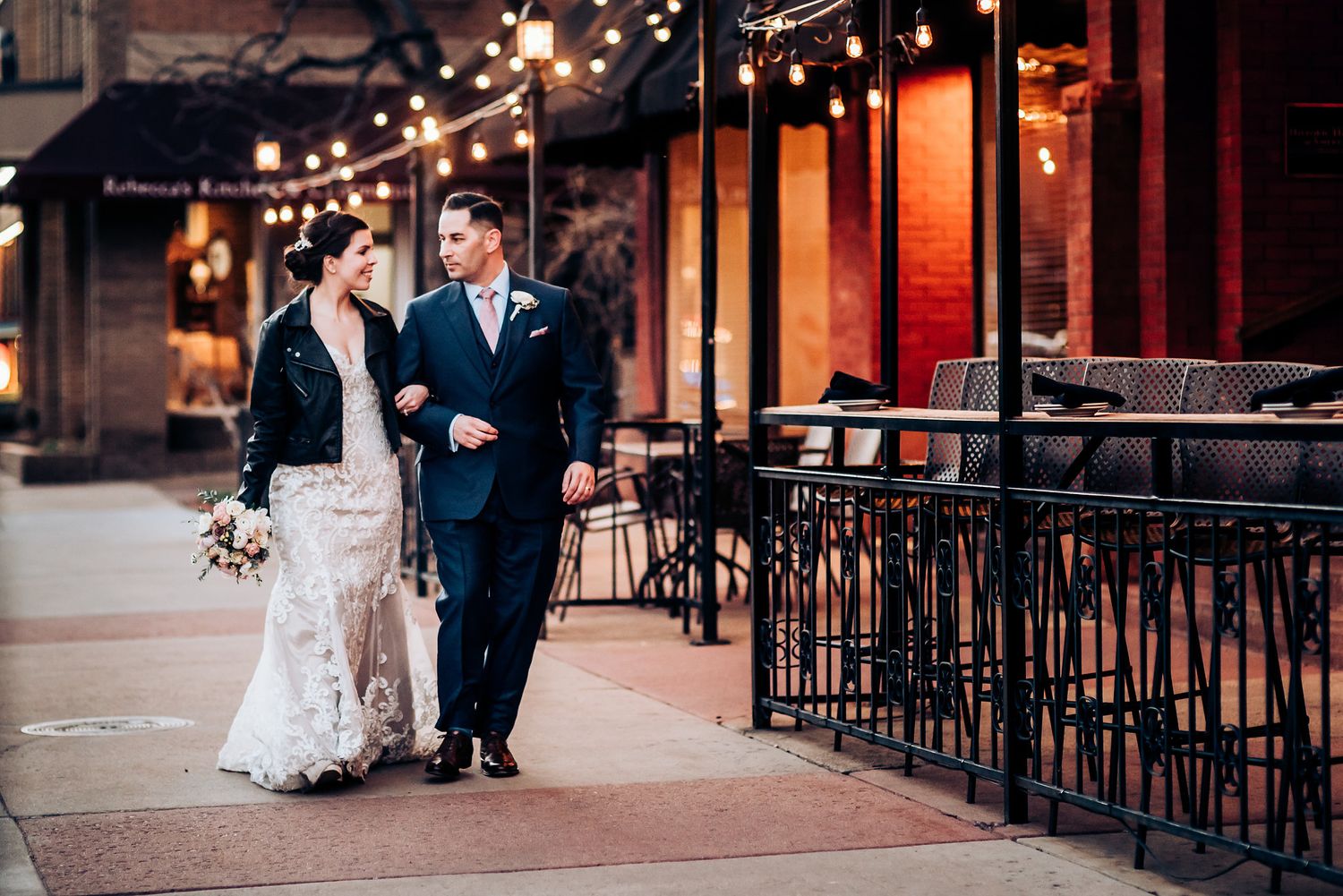 Wedding couple walks along restaurant patio with string lights and iron railings.