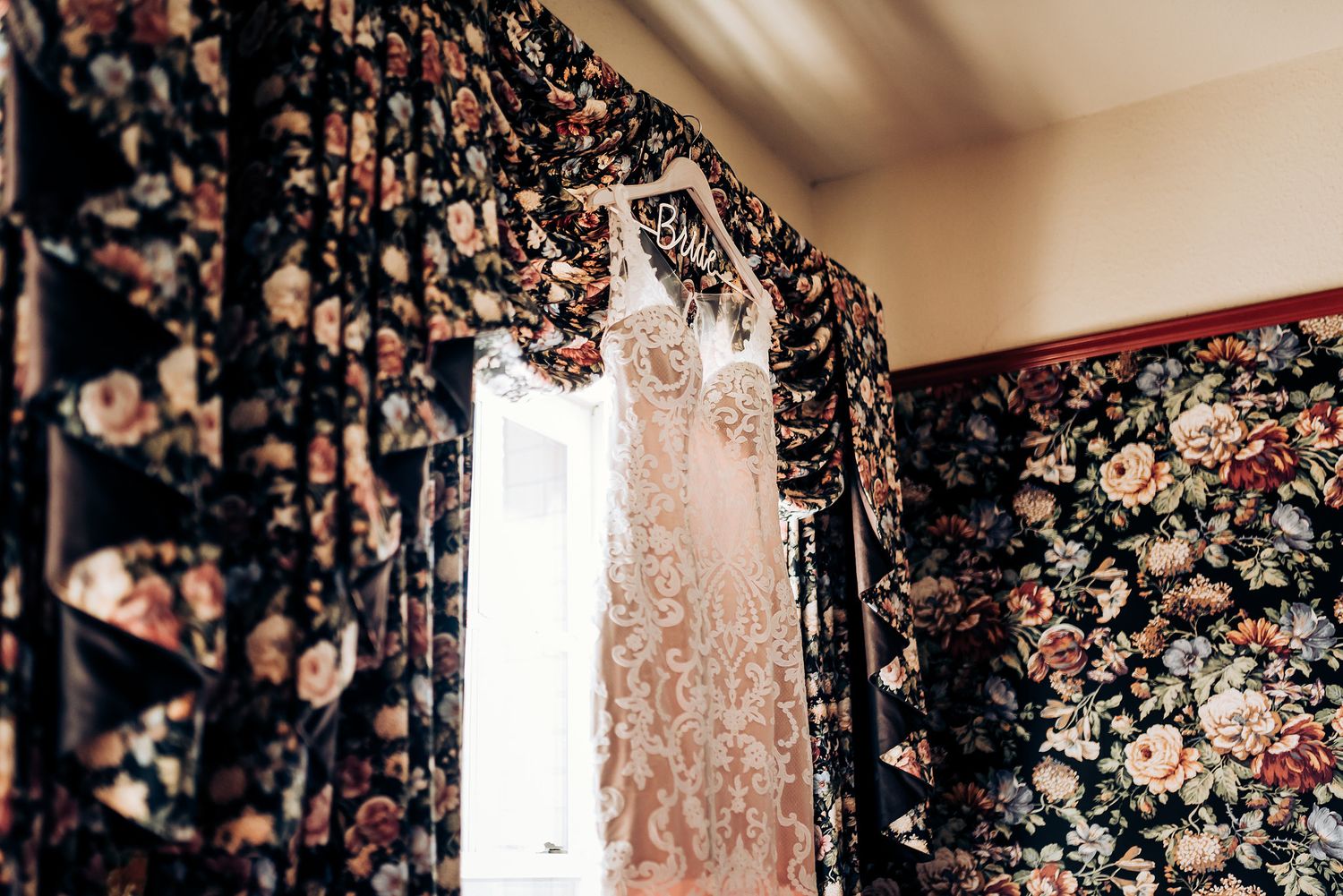 Elegant white wedding dress hanging against dark floral wallpaper with window light.