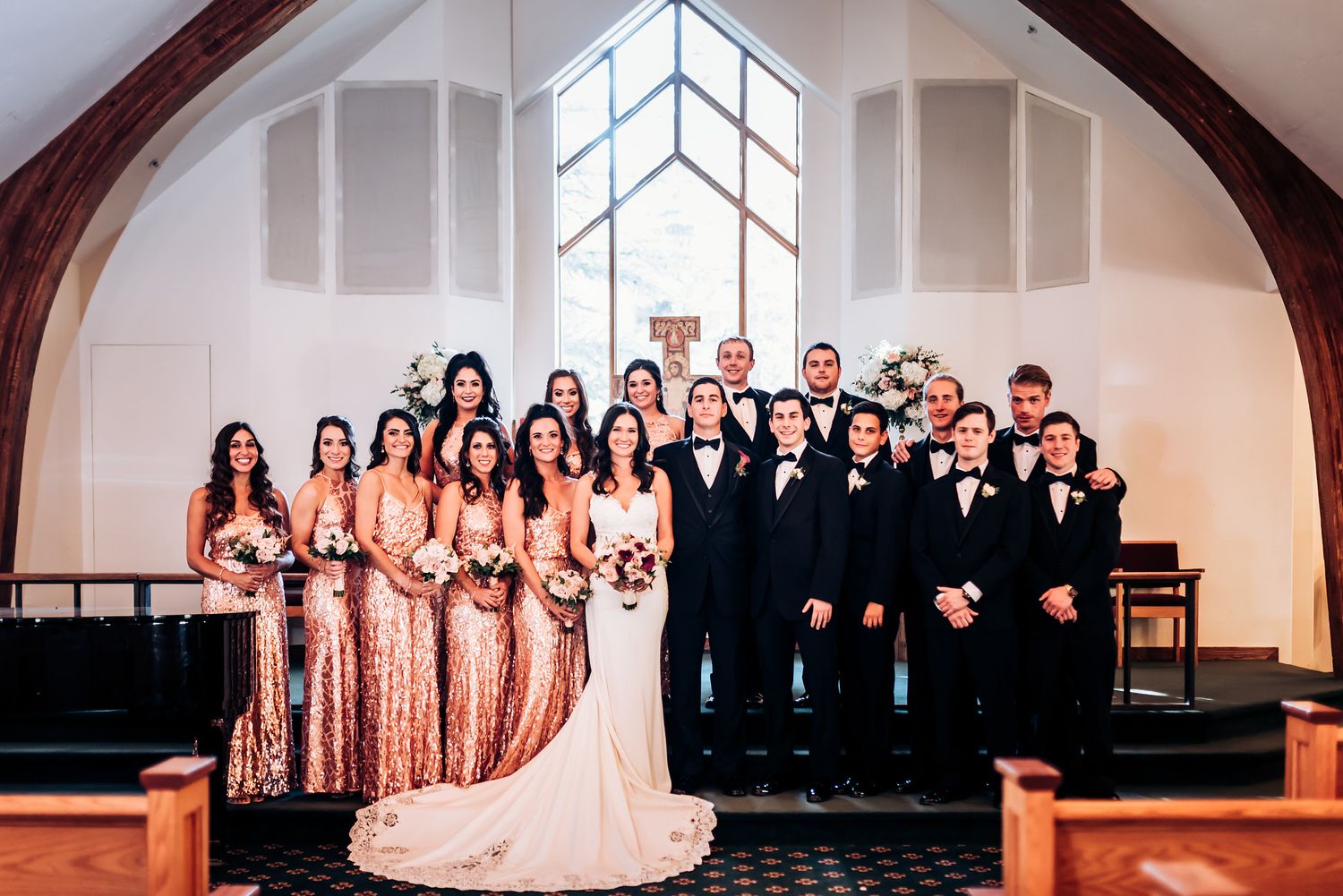 Wedding party poses in church with bridesmaids in rose gold dresses and groomsmen in black suits.