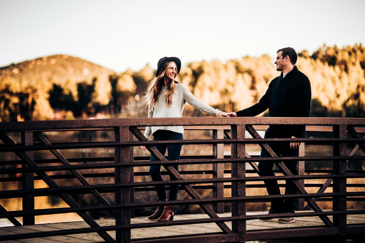 Couple standing together on wooden bridge at sunset in mountainous landscape.