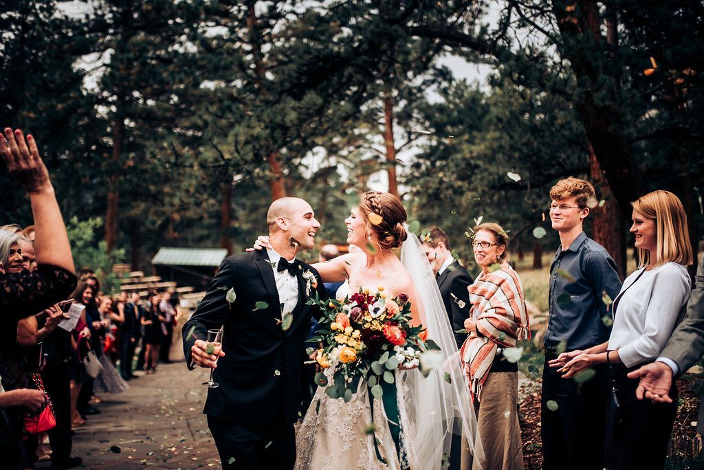 Wedding party walks down an outdoor aisle lined with guests in a pine forest setting.