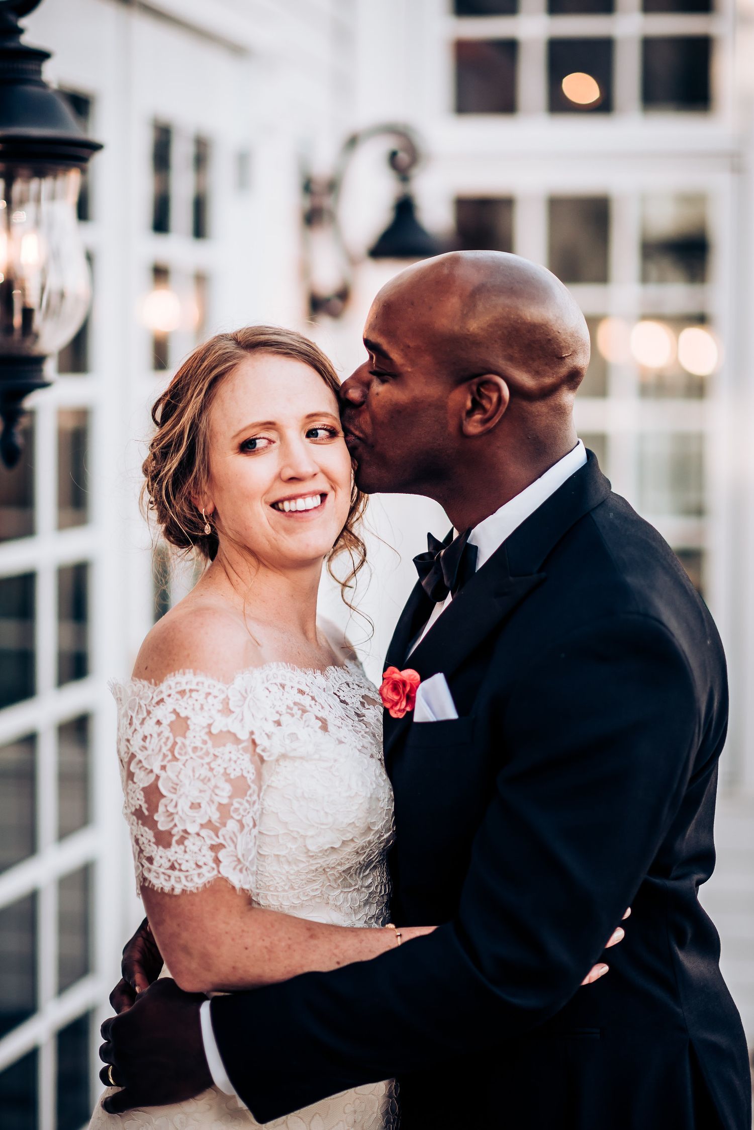 Loving wedding portrait of couple embracing outdoors with white architecture in background.