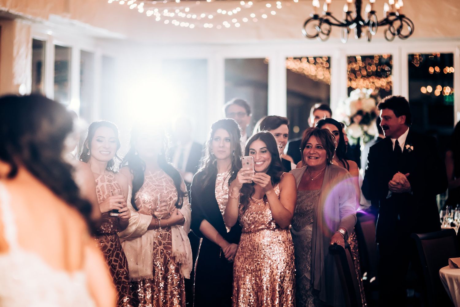 Wedding guests in sparkly rose gold dresses celebrate during reception under string lights.