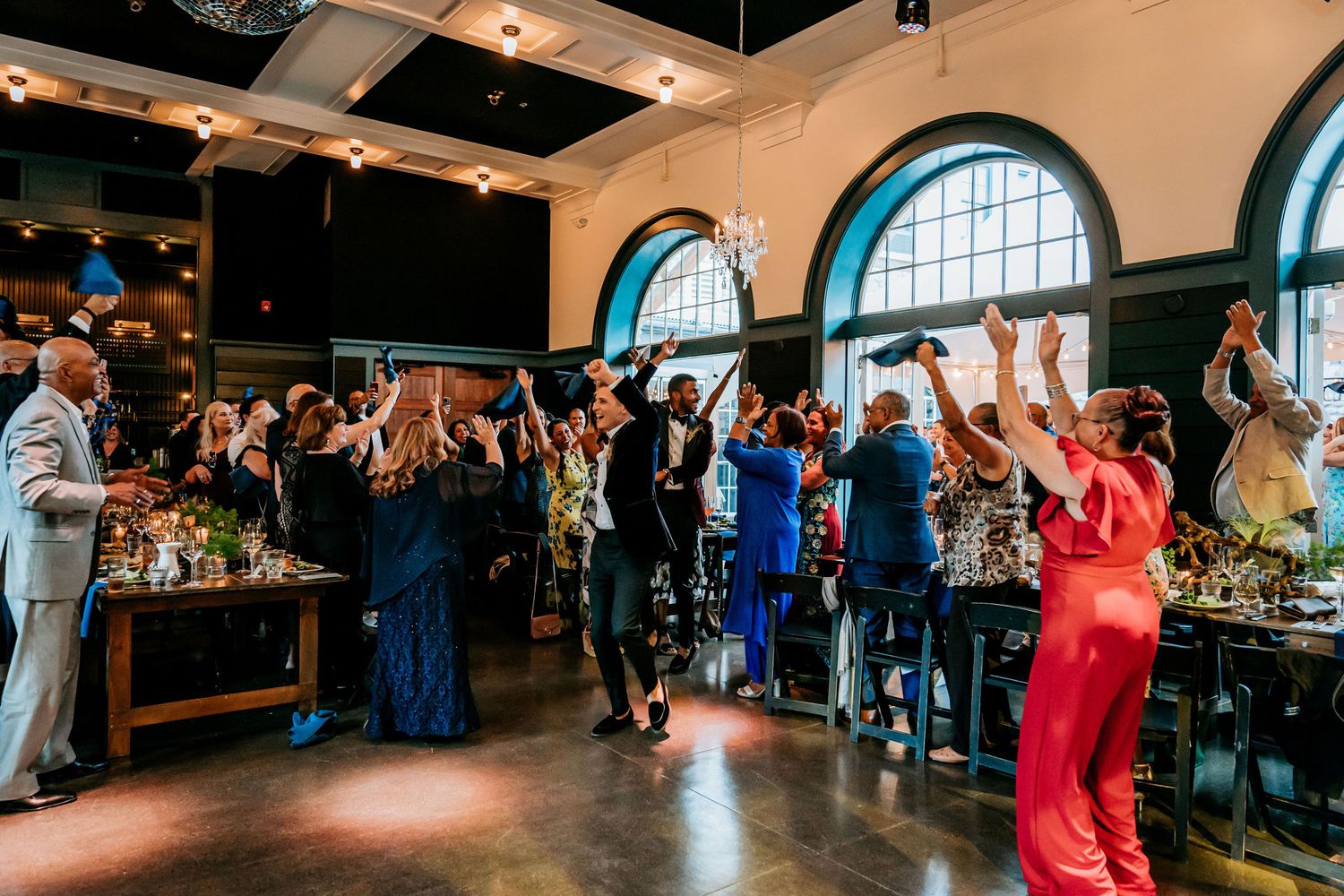 Wedding guests dancing and celebrating in an elegant venue with arched windows.