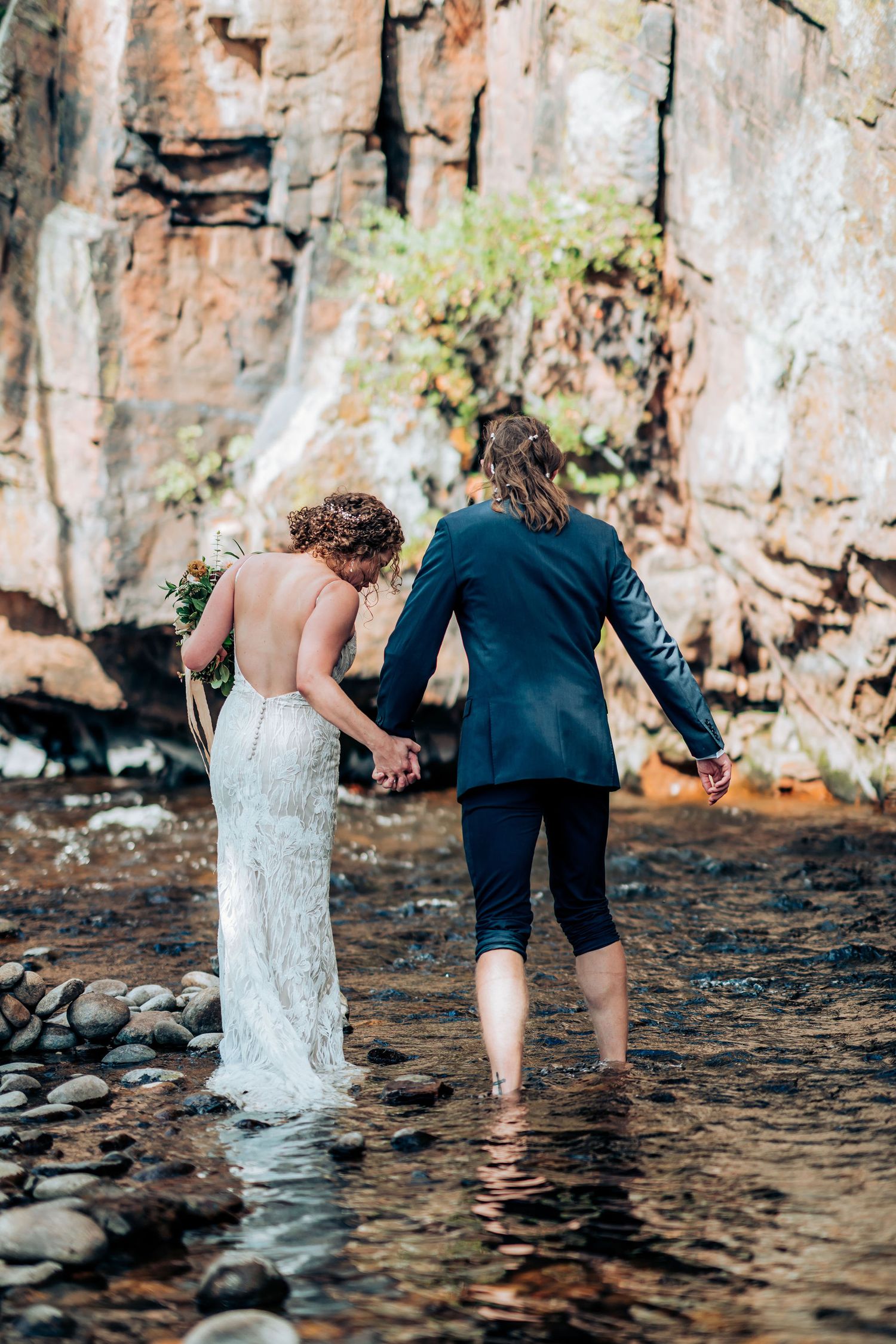 A couple wades together in shallow water near a rocky cliff face during their wedding photoshoot.