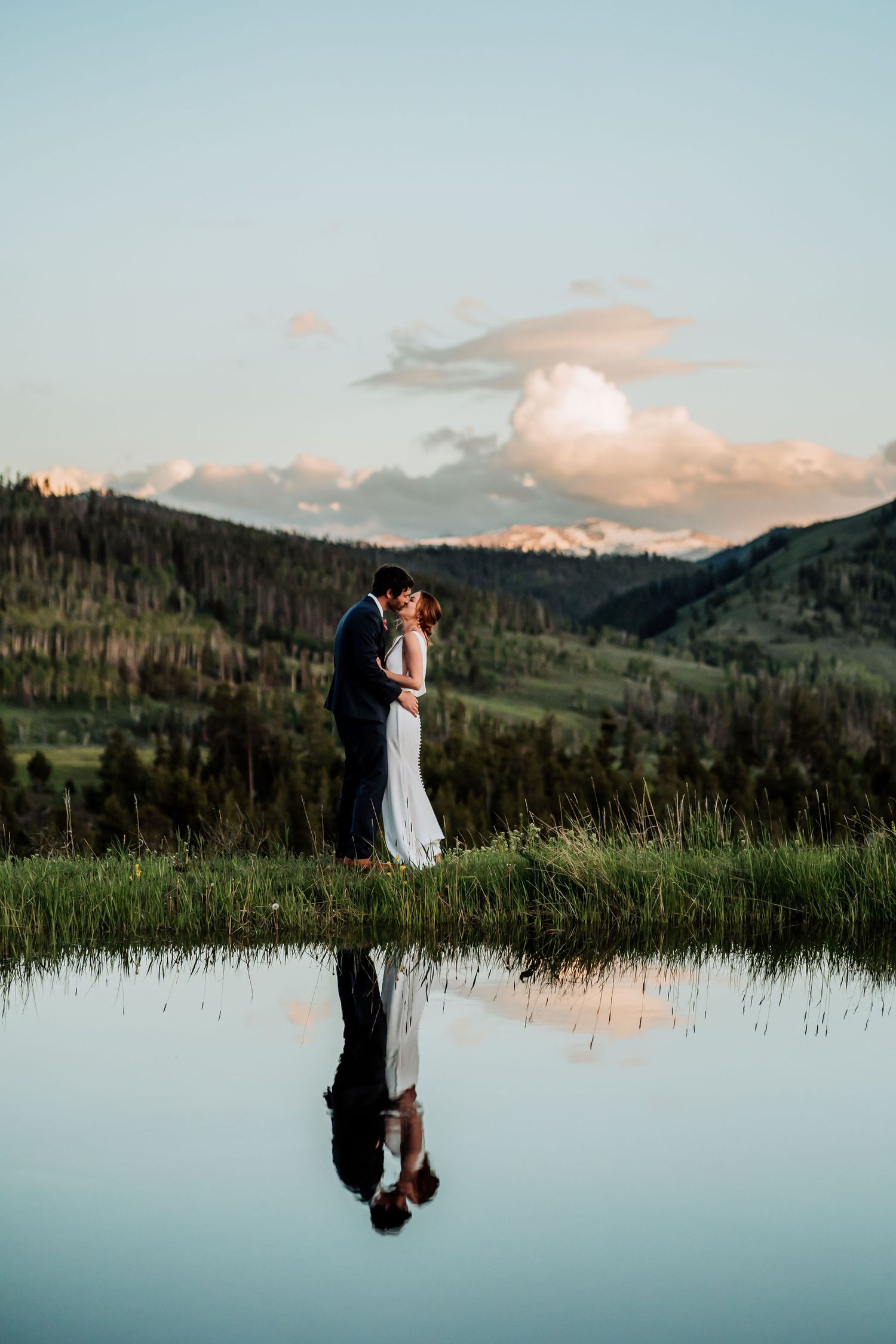 Wedding couple embraces by mountain lake during golden hour with perfect reflection.