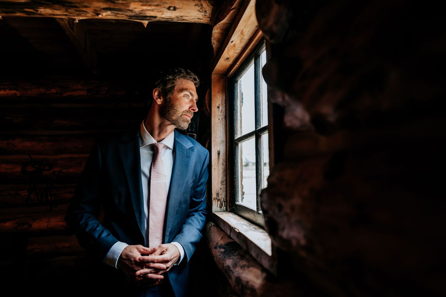 Person in blue suit standing by window in rustic log cabin setting.