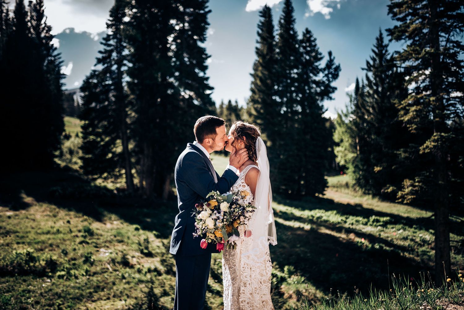 Newlyweds embrace in mountain meadow with evergreen trees and dramatic sky backdrop.