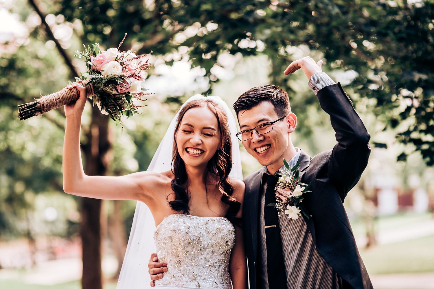 A joyful wedding couple celebrating outdoors with raised arms and pink bouquet.