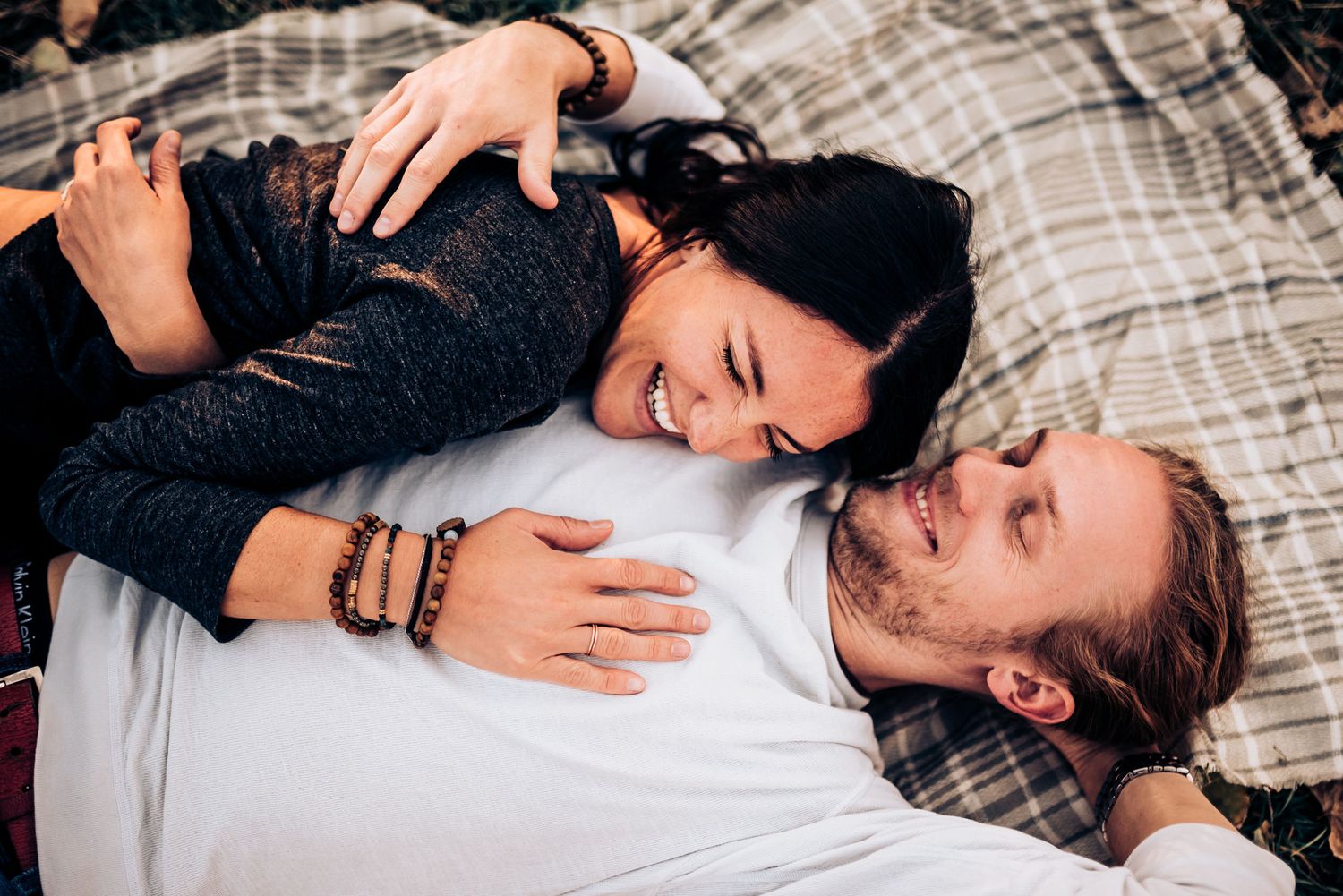 A couple giggling and embracing while lying on a plaid blanket outside.